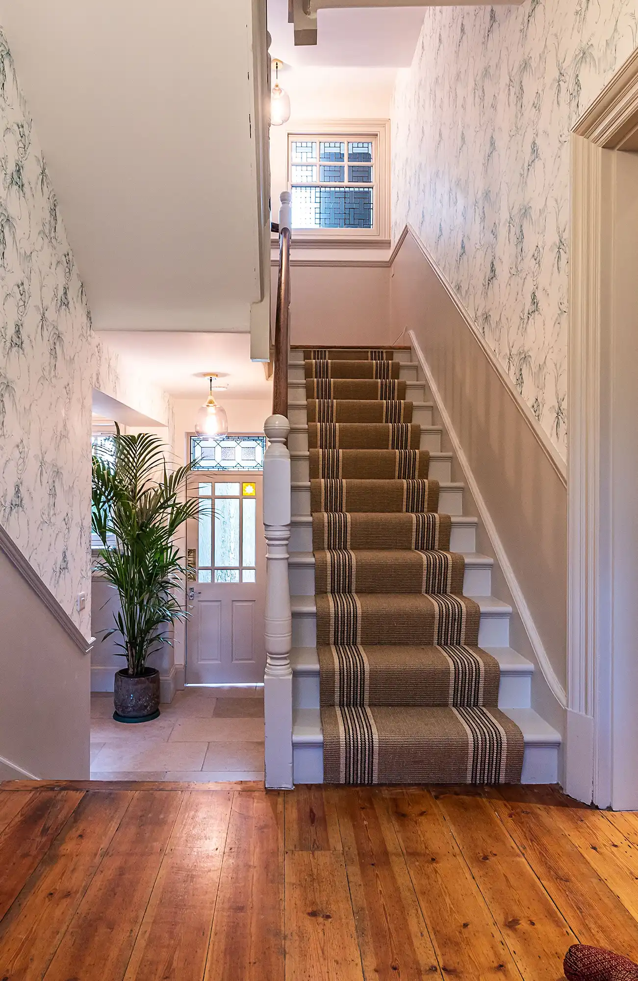 Bright entryway with a staircase, patterned carpet, potted plant, and a decorative door leading outside.