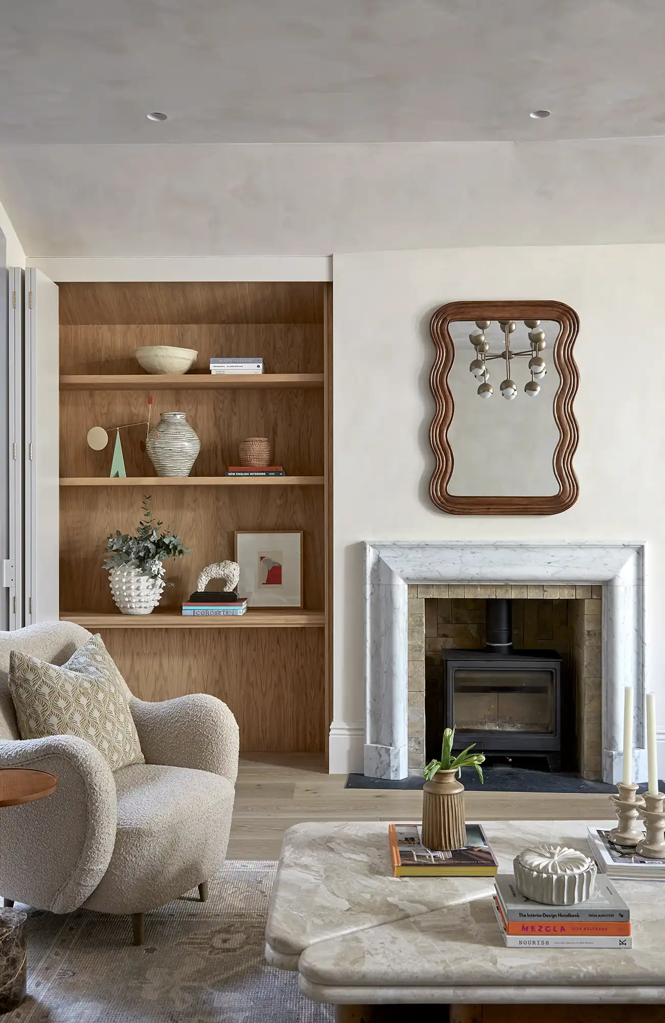 Modern living room featuring a cozy chair, elegant marble fireplace, open shelves with decor, and a stylish mirror.