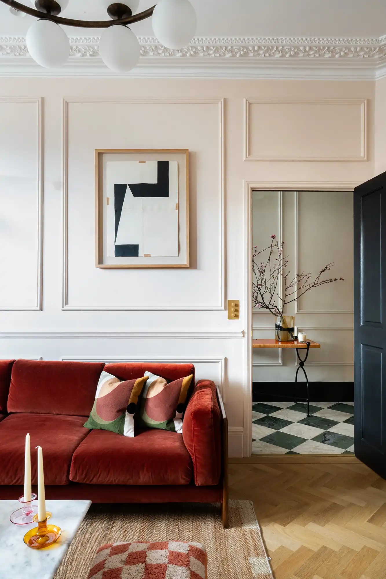 Cozy living room featuring a red velvet sofa, patterned pillows, and an abstract artwork on the wall. A doorway leads to another room.